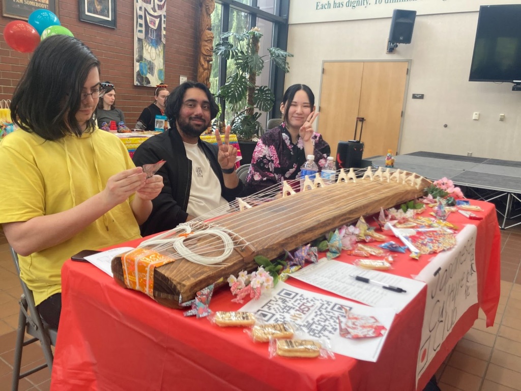 In an auditorium. Three students sit at a table with a red table cloth. There are candies all over the table, as well as a sign up sheet and full page QR code. A large wooden instrument with strings that is the length of the table, called a Koto, sits atop the candies. 