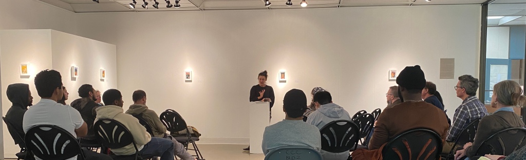 A square room with white walls. Art is hanging in a perpendicular column at the center of the wall. A woman stands at a podium where a book is placed. She is reading from the book to a crowd. 