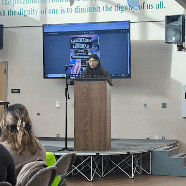 A women stands on a stage reading from a podium into a microphone. There is a tv behind displaying the cover of the book Living in Two languages. 
